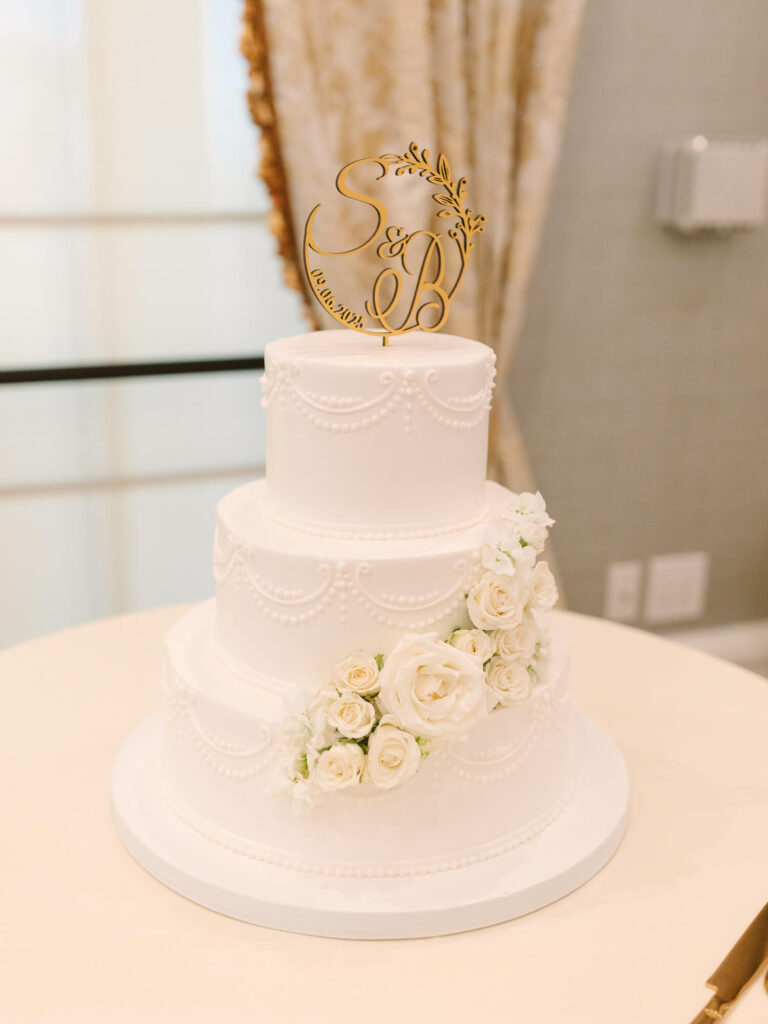 Three-tiered white wedding cake adorned with pearl-like icing patterns and white roses. A gold monogram topper adds elegance. 
