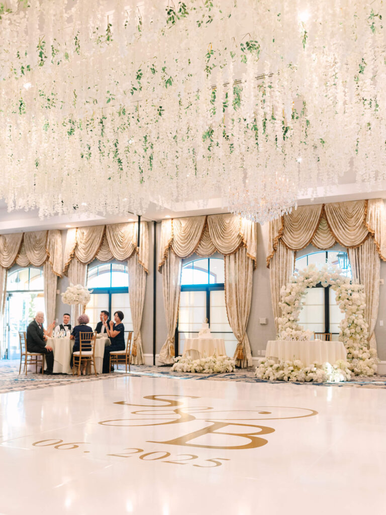 Elegant ballroom with cascading white floral ceiling decor, gold drapes, round tables with guests, and a floral arch. Dance floor features initials and date.
