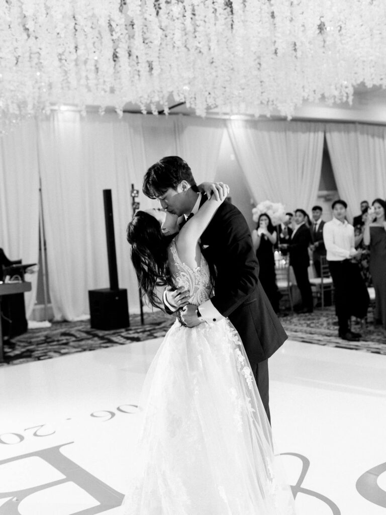 Black and white image of a couple sharing a kiss during their wedding dance. The bride wears a flowing gown; guests watch with joy in the softly-lit venue.