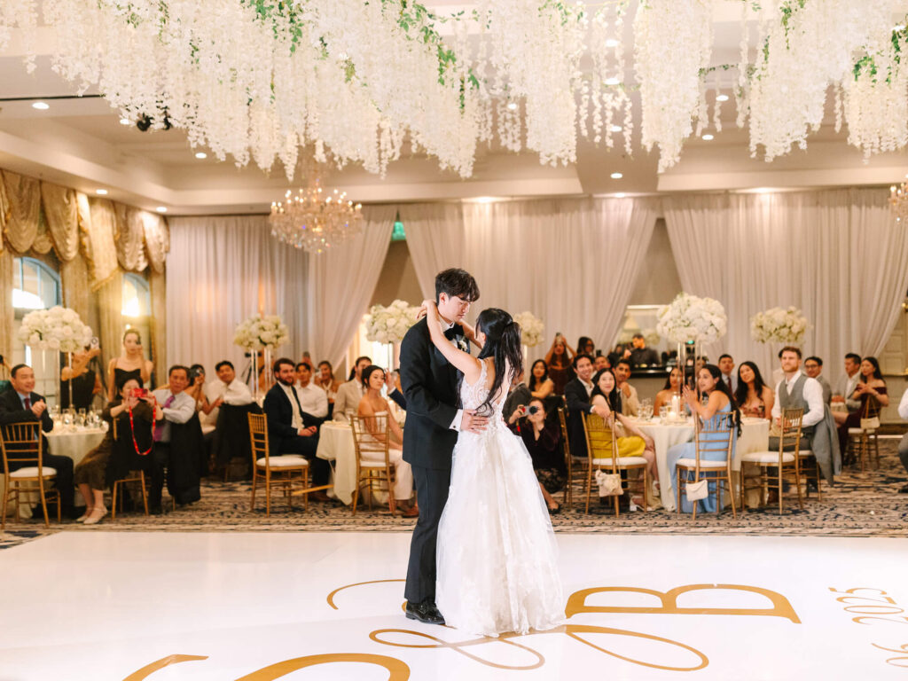 A couple shares a romantic dance at their wedding, surrounded by elegantly dressed guests seated at tables. The venue features white floral decorations and soft lighting.