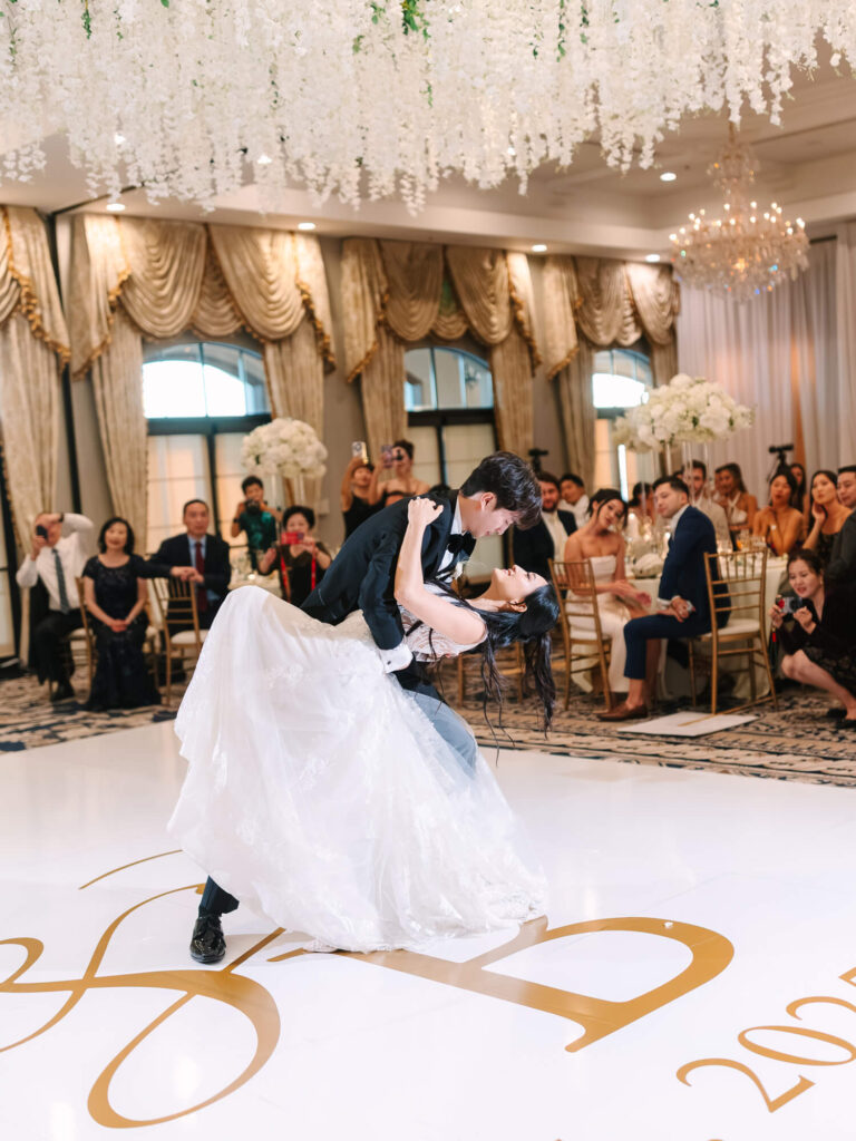 A couple shares a romantic dance dip at their wedding, surrounded by guests seated in a lavish venue with golden drapes and hanging floral decor.