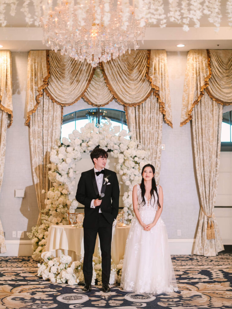 A couple stands smiling in wedding attire; the groom in a black tuxedo, the bride in a white gown. They're framed by elegant curtains and white floral arrangements, with a grand chandelier above.