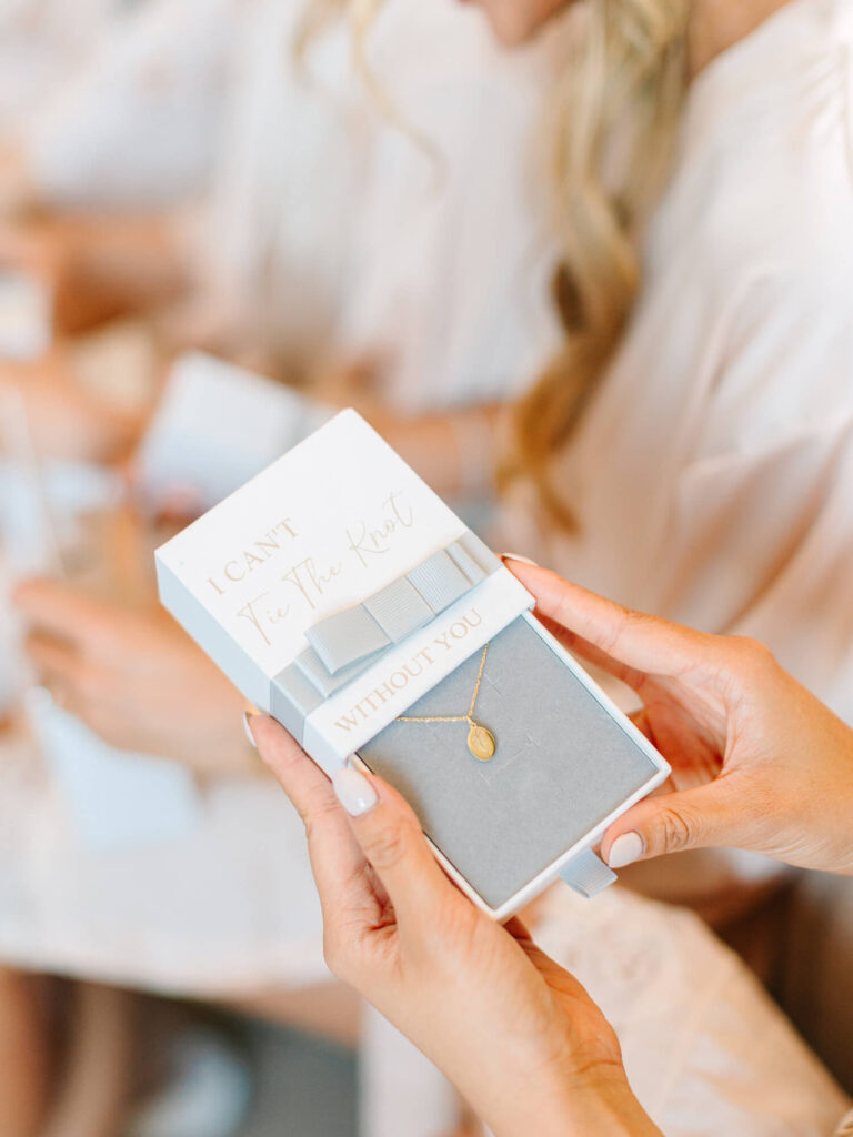 A close-up of hands holding a gift box with a gold necklace inside. The box reads "I Can't Tie the Knot Without You," conveying a heartfelt message.