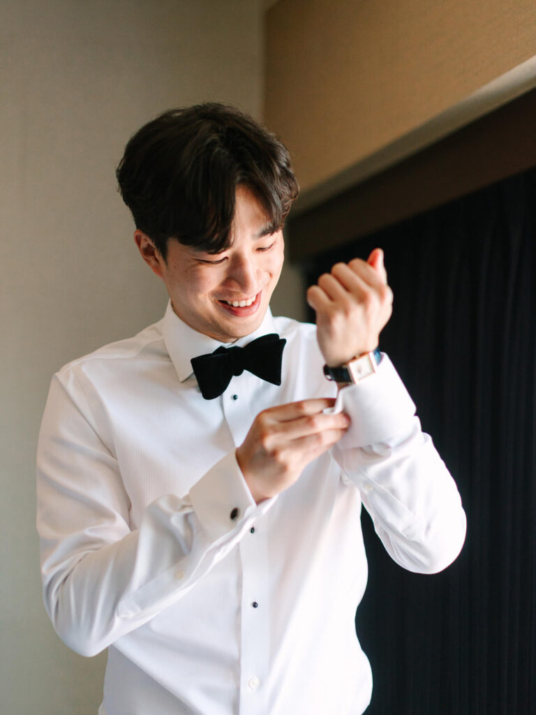 Young man in formal attire smiles while adjusting his watch. He's wearing a white shirt and black bow tie.