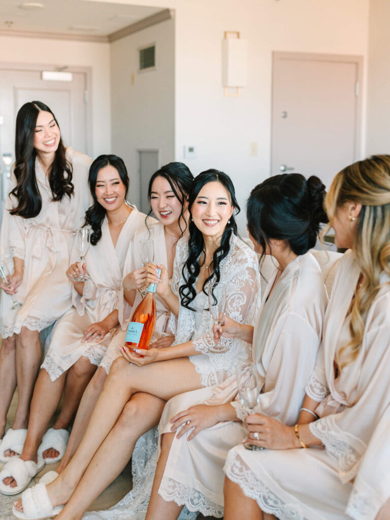 Bride and bridesmaids in matching robes sit joyfully on a couch, holding champagne glasses. The bride holds a bottle.
