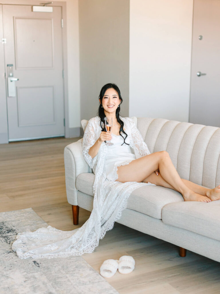 A woman in a white lace robe smiles and relaxes on a light gray sofa, holding a champagne glass. Her slippers rest on a patterned rug.