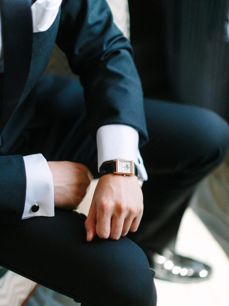 A person in a sleek black suit sits, showing sophistication. Focus on a rectangular watch and cufflinks