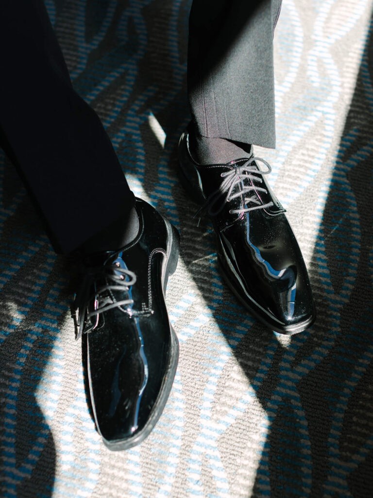 A person wearing polished black dress shoes stands on a patterned carpet, with light casting sharp shadows.