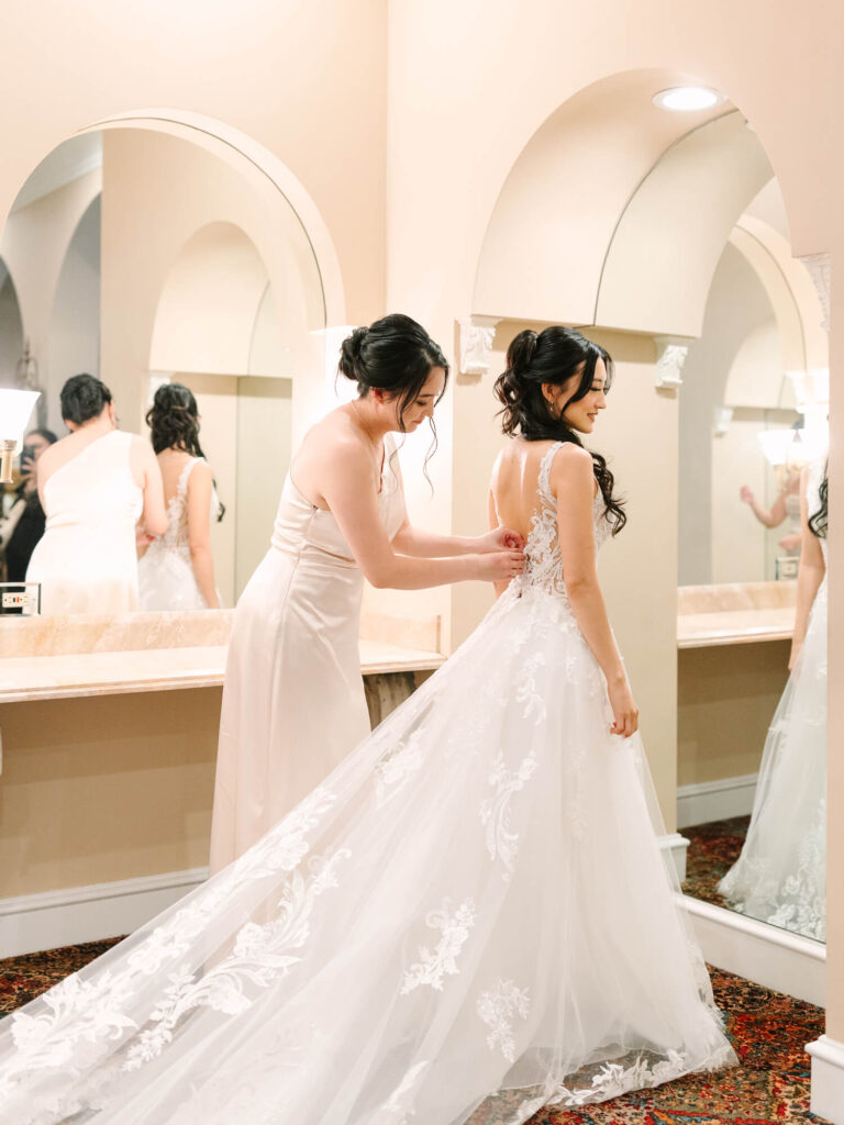 A woman helps another with her lace wedding dress in a warmly lit room with large mirrors