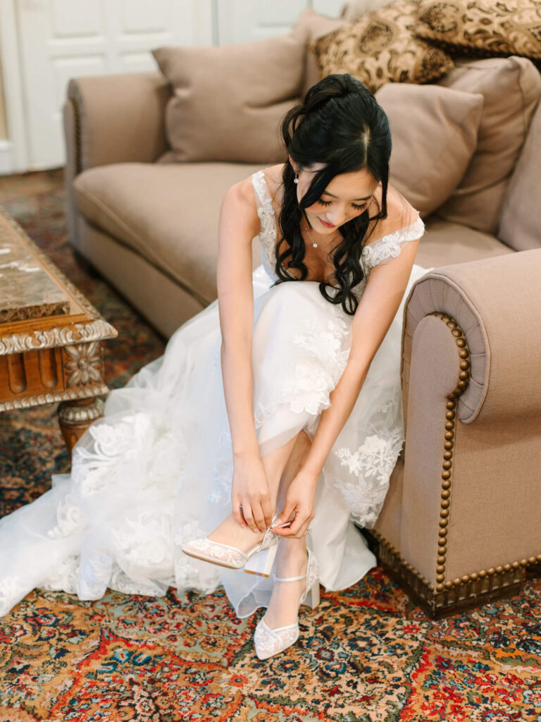 Bride sitting on a beige sofa, wearing a white wedding dress with lace details. She adjusts her lace shoes.