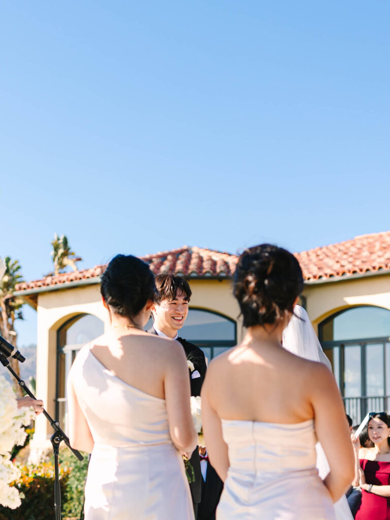 A groom smiles at his bride during an outdoor wedding, under a clear blue sky. Bridesmaids in pale dresses stand nearby; a tile-roofed building is behind them.