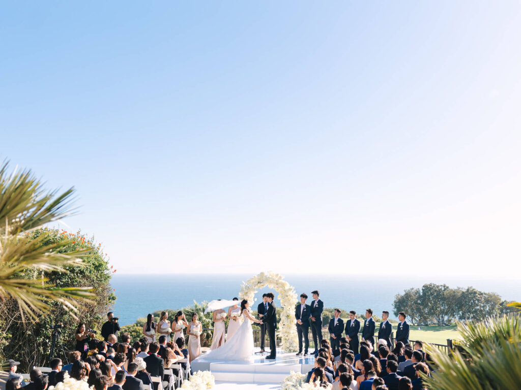 Outdoor wedding ceremony overlooking the ocean. A couple stands under a floral arch, surrounded by bridesmaids and groomsmen, with guests seated nearby.