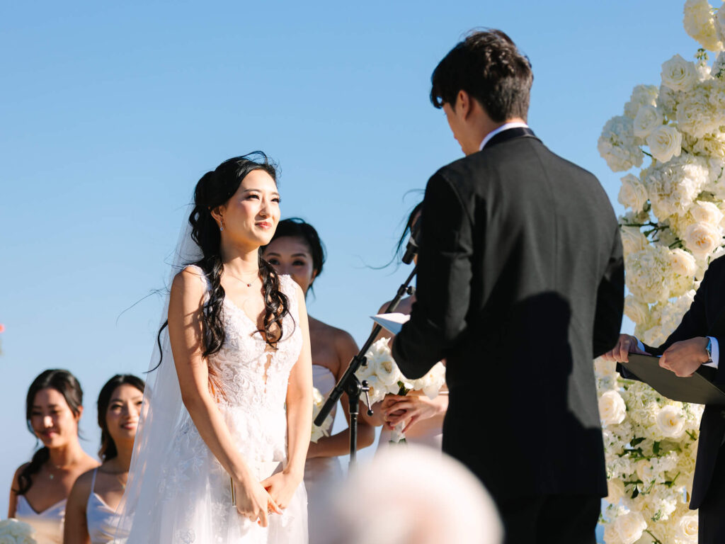 A bride in a white dress smiles warmly at a groom in a black suit during an outdoor wedding ceremony. Bridesmaids in light dresses and floral arrangements are in the background, with a clear blue sky.