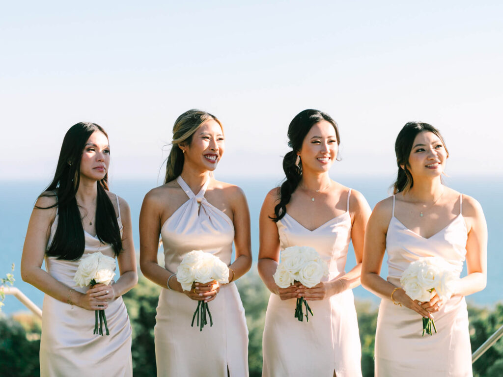 Four women in matching cream dresses stand outdoors, holding white rose bouquet.