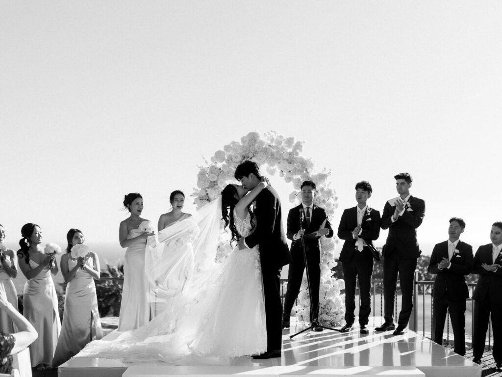 A bride and groom kiss under a floral arch, surrounded by a cheering wedding party.