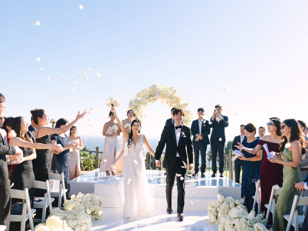 A joyful bride and groom walk hand in hand down the aisle outside, surrounded by guests tossing flower petals. A floral arch and bright skies frame the scene.