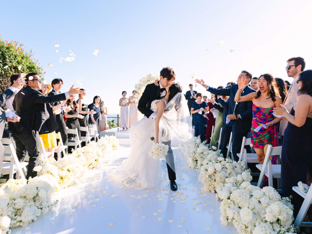 Bride and groom kiss on a flower-lined aisle, surrounded by cheering guests throwing petals.