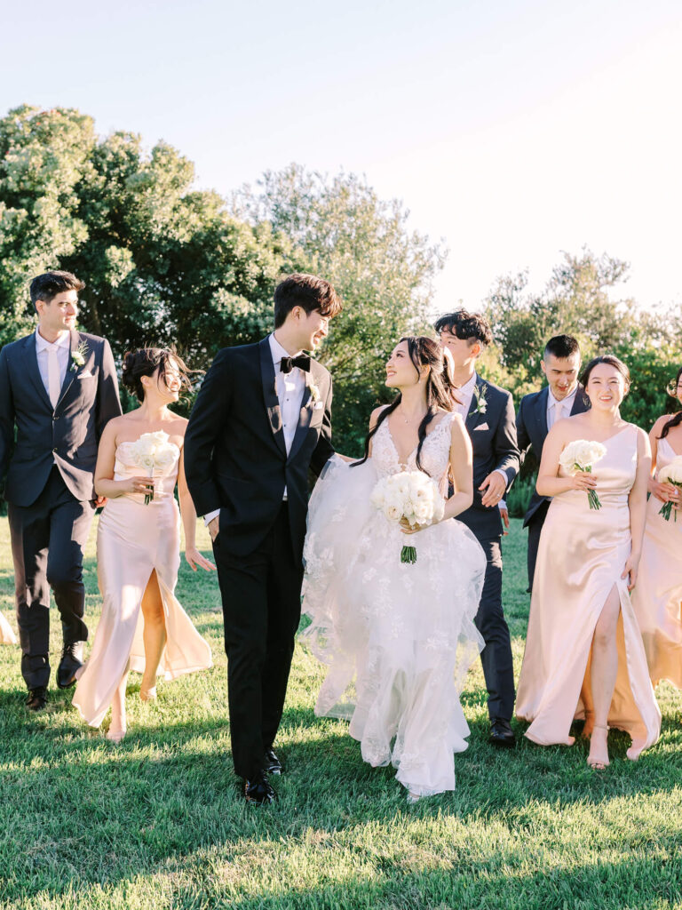 A joyful wedding party walks on grass. The bride in a white gown holds a bouquet, smiles at the groom in a black tuxedo. Bridesmaids in light dresses and groomsmen in suits share a happy moment under a sunny sky with lush trees.