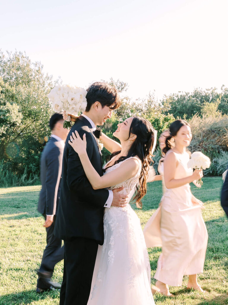 A bride and groom embrace joyfully outdoors. The bride wears a white gown, the groom a black suit. Bridesmaids with bouquets walk by, all under sunny skies and surrounded by greenery.
