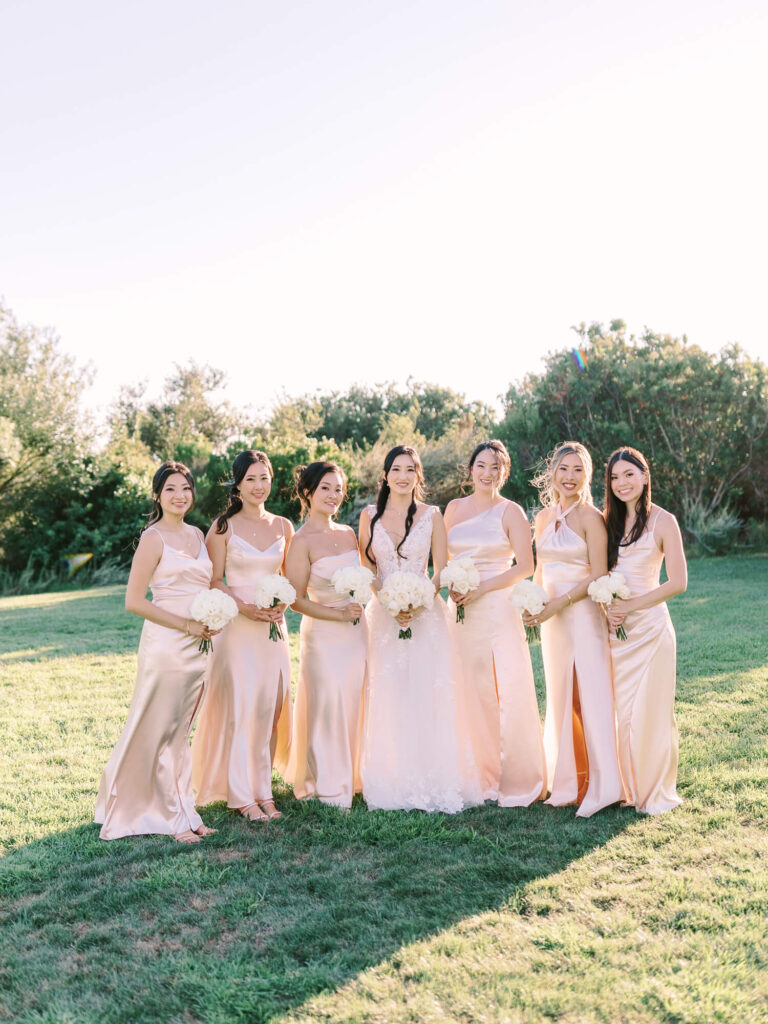 Bride and six bridesmaids in soft pink dresses, holding white bouquets, stand smiling on grass with trees in background under a clear sky.