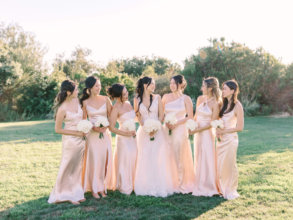 A bride in a white gown stands with six bridesmaids in blush dresses, holding white bouquets. They smile at each other.