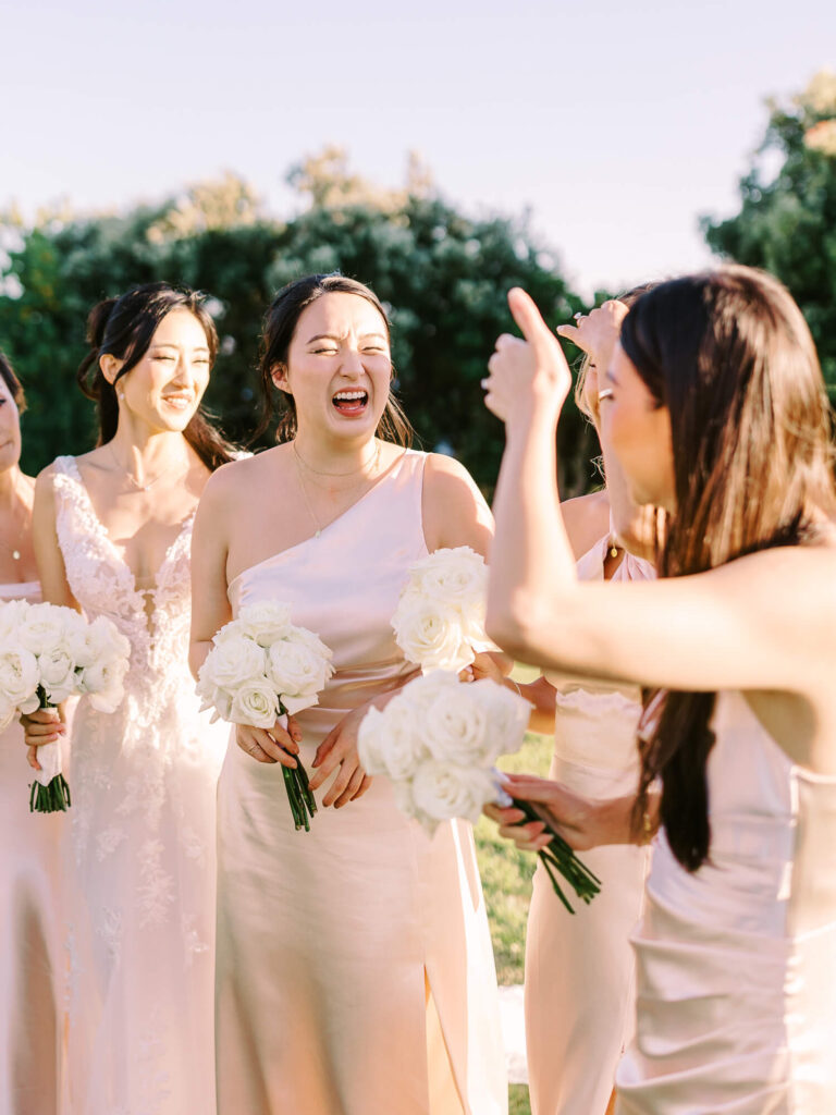 Bridesmaids in pale pink dresses laugh and chat outdoors, holding white rose bouquets.