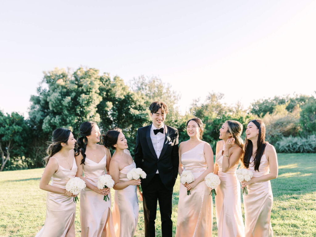 A group of smiling bridesmaids in light pink dresses stand with a groomsman in a black tuxedo on a sunny lawn, holding white bouquets.