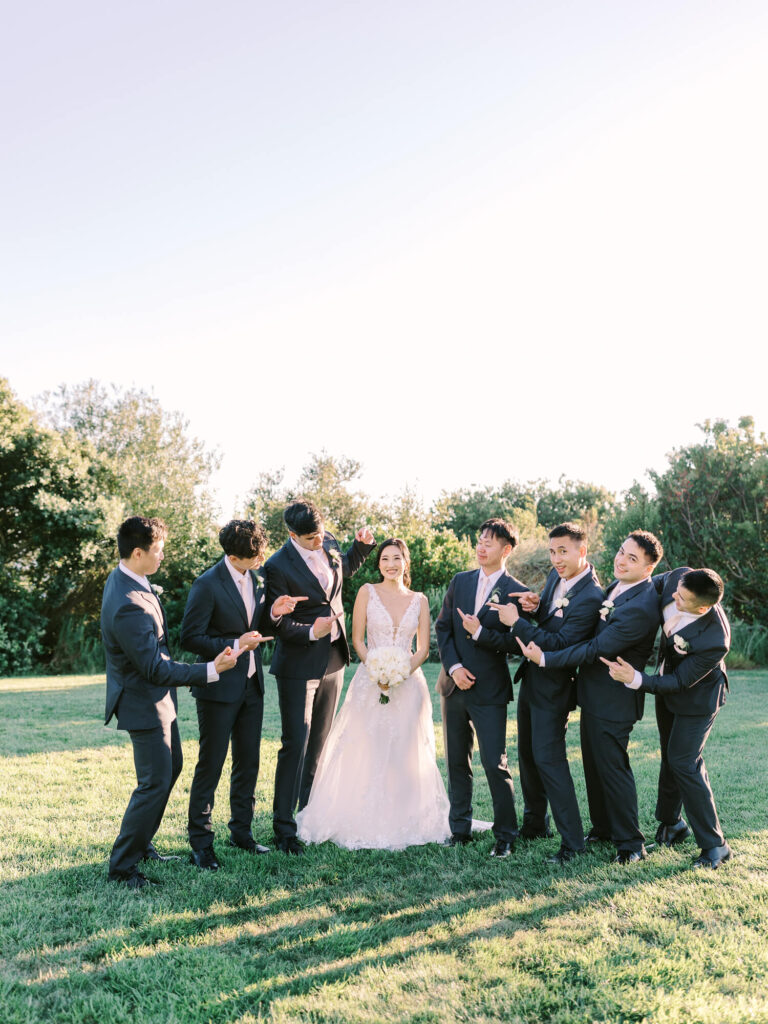Bride in white gown smiles amid groomsmen in blue suits on a sunny lawn. They point playfully at her.