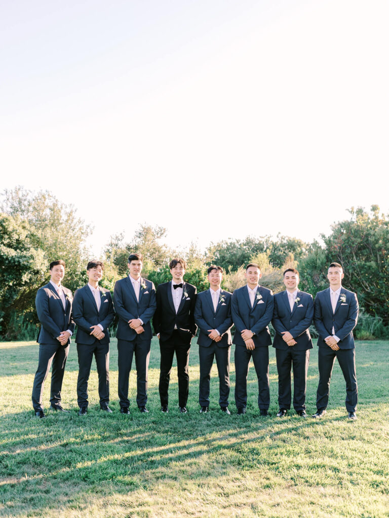 A group of eight men stand on grass in sunlight, seven in matching gray suits, one in a black suit with a bow tie. 