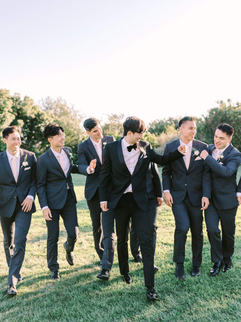 Seven men in suits walk across a grassy field, smiling and laughing. The groom is centered, wearing a bow tie.