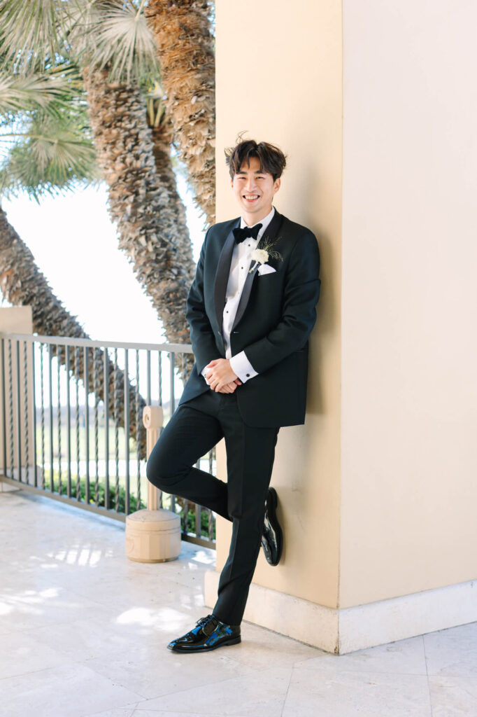 Young man in a tuxedo smiles while leaning against a beige wall, with palm trees and a bright, sunny background.
