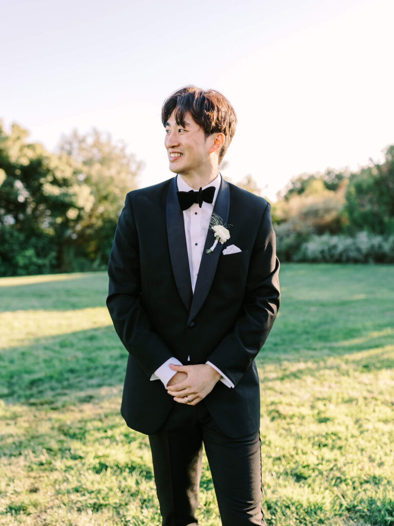 Young man in a tuxedo stands smiling on a sunny lawn, hands folded. Green trees and bushes form a backdrop.