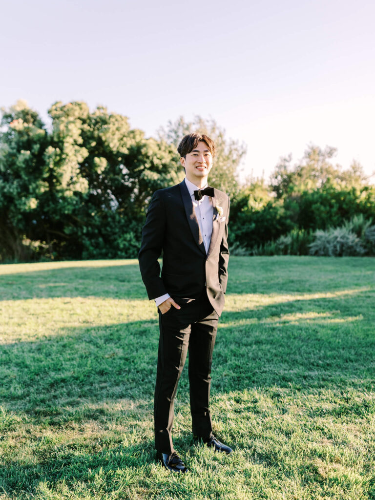 A man in a black tuxedo and bow tie stands on a sunlit grassy lawn, smiling with one hand in his pocket.