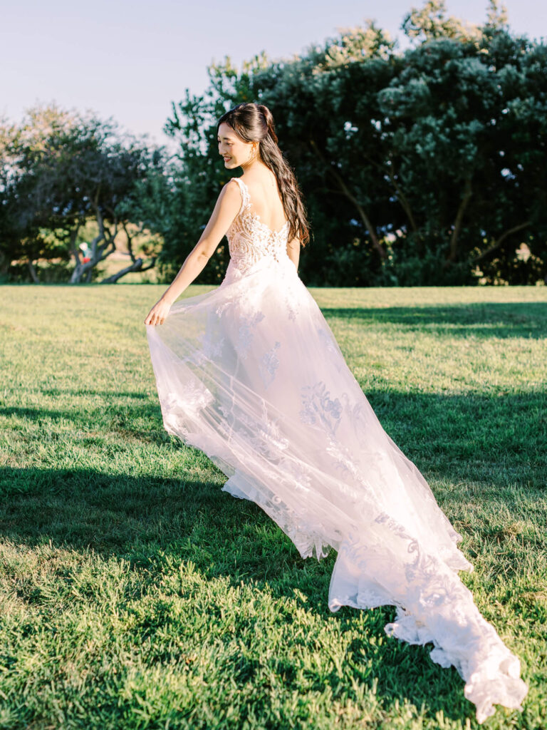 A woman in a flowing white wedding dress with lace details walks on a sunlit grassy field, surrounded by trees.