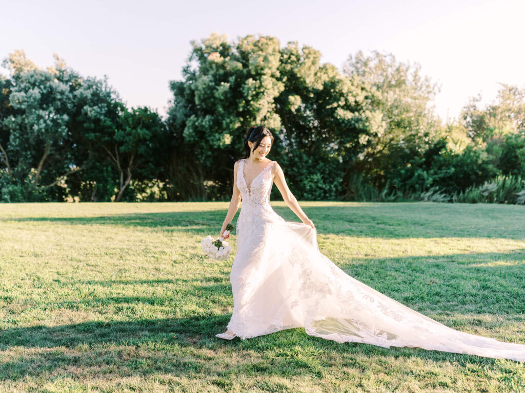 A bride in a flowing white gown holds a bouquet, standing on a sunlit grass field.