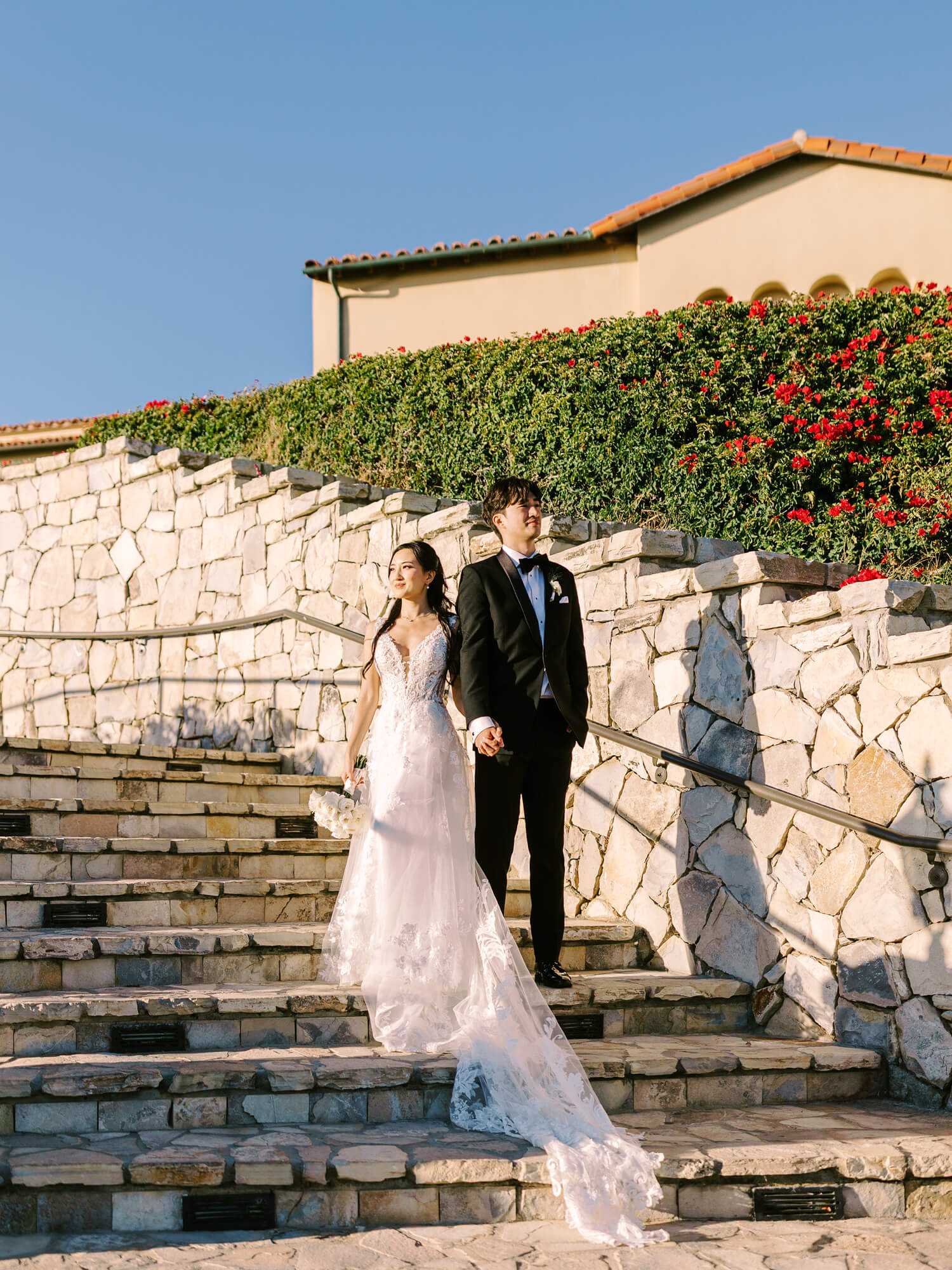 A bride in a flowing white gown and a groom in a black tuxedo walk hand-in-hand down stone steps. The background shows a building with red-tiled roofs and vibrant greenery under a clear blue sky, conveying a joyful wedding atmosphere.