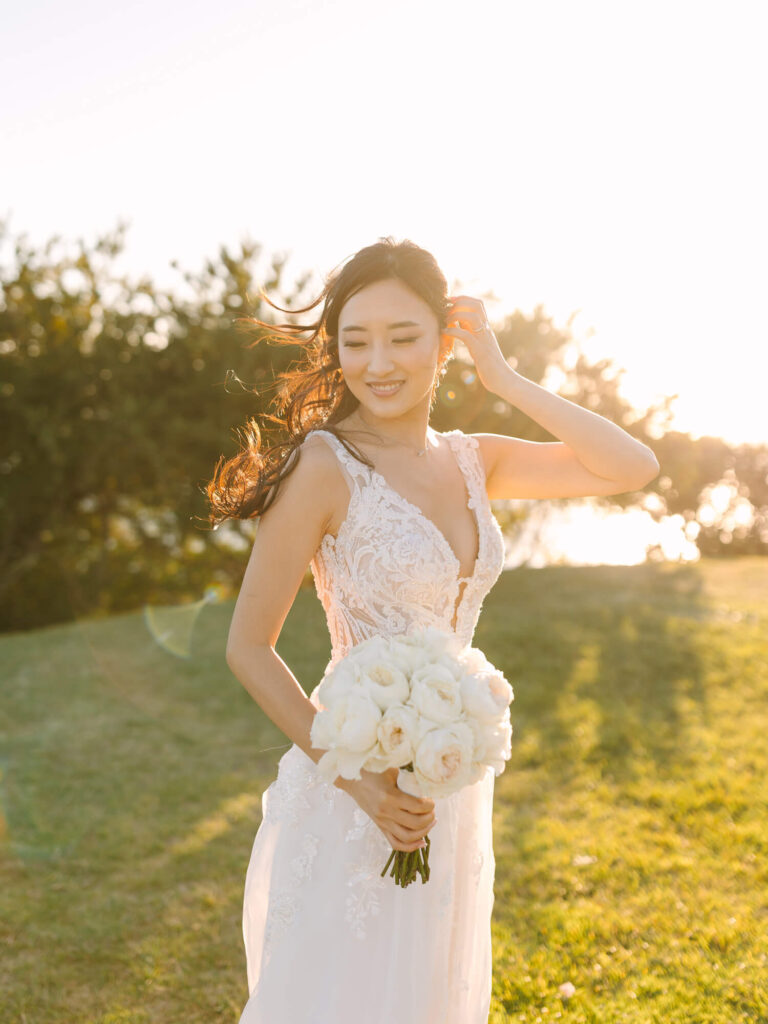 A bride in a lace wedding dress smiles joyfully, holding white roses. Sunlight illuminates her hair.