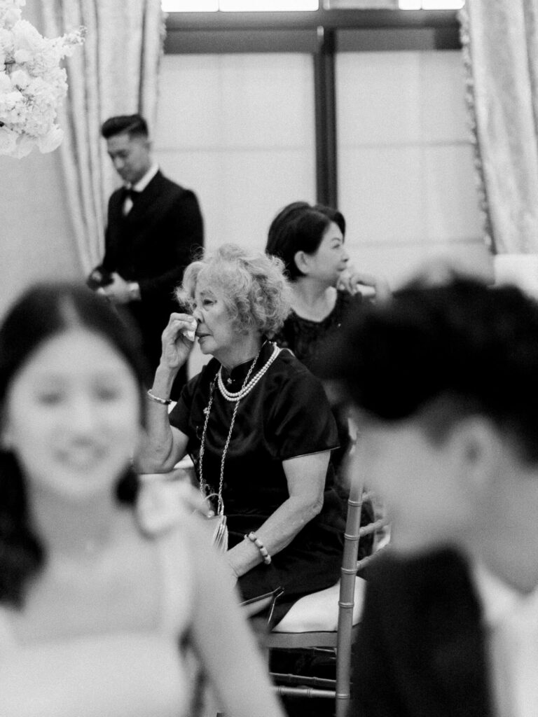 A black-and-white photo of an elderly woman in a dark dress, sitting and wiping her eye emotionally at an elegant event.