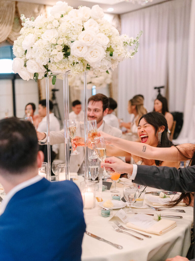 Guests cheerfully toast around a table adorned with tall white floral arrangements.