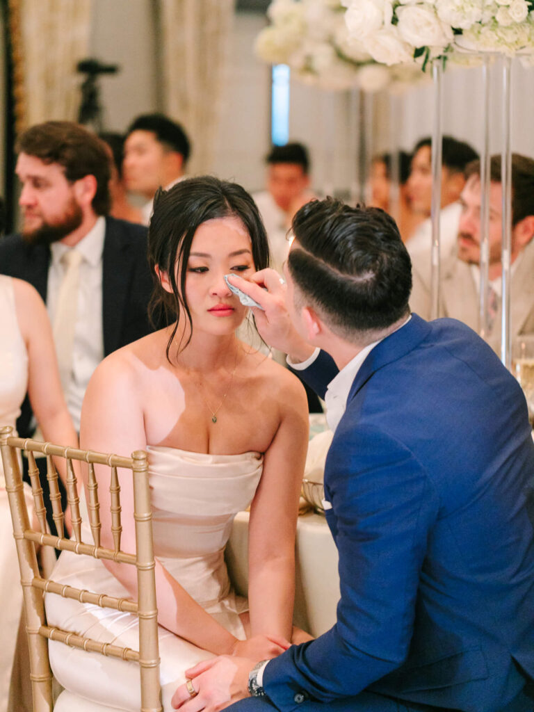 A man in a blue suit gently wipes tears from a woman's face at a wedding reception.