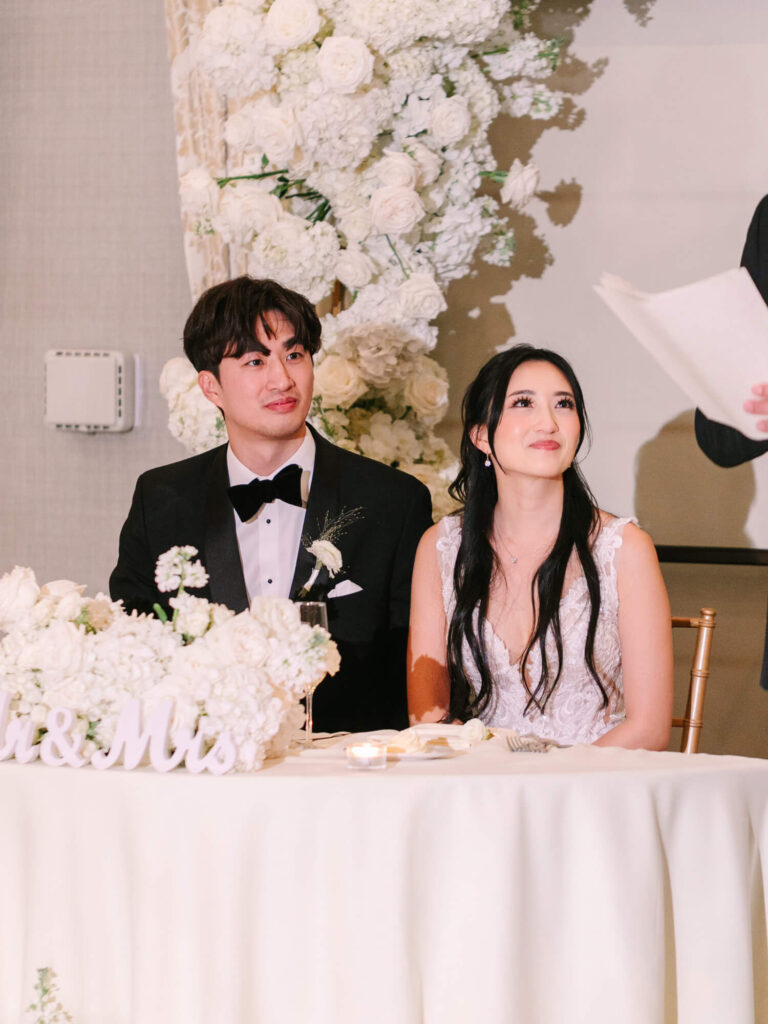 A bride and groom sit at their wedding table, adorned with white flowers and a "Mr. & Mrs." sign. They look attentive and content. Floral backdrop.