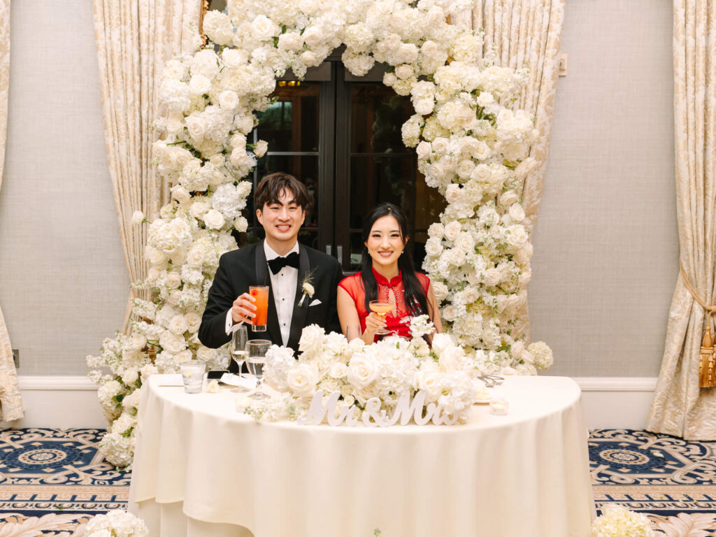 A couple sitting at a white-clothed table, smiling and holding drinks. They're framed by a large arch of white flowers.