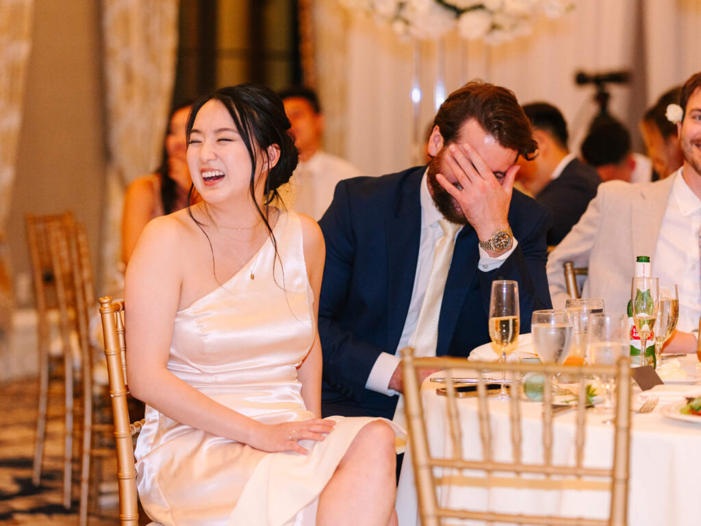 A woman in a satin dress laughs heartily, while a man in a blue suit covers his face in amusement at a wedding reception.