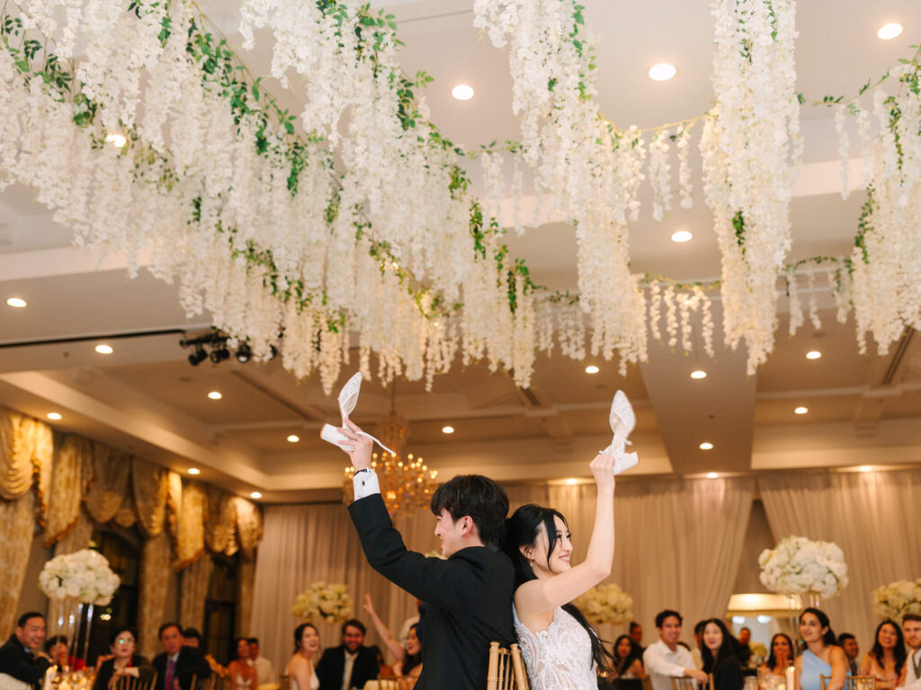 A couple sits back-to-back holding shoes in a wedding hall with guests around them. White flowers hang from the ceiling.