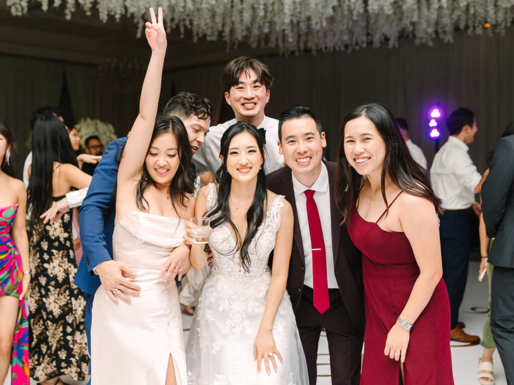 A joyful group poses at a wedding reception. The bride in a white dress smiles at the center, surrounded by friends in formal attire.