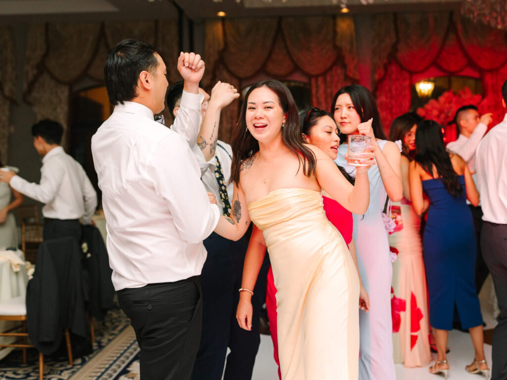 A joyful group dances at a lively party. A woman in a yellow dress smiles and raises a drink, surrounded by others in colorful outfits.