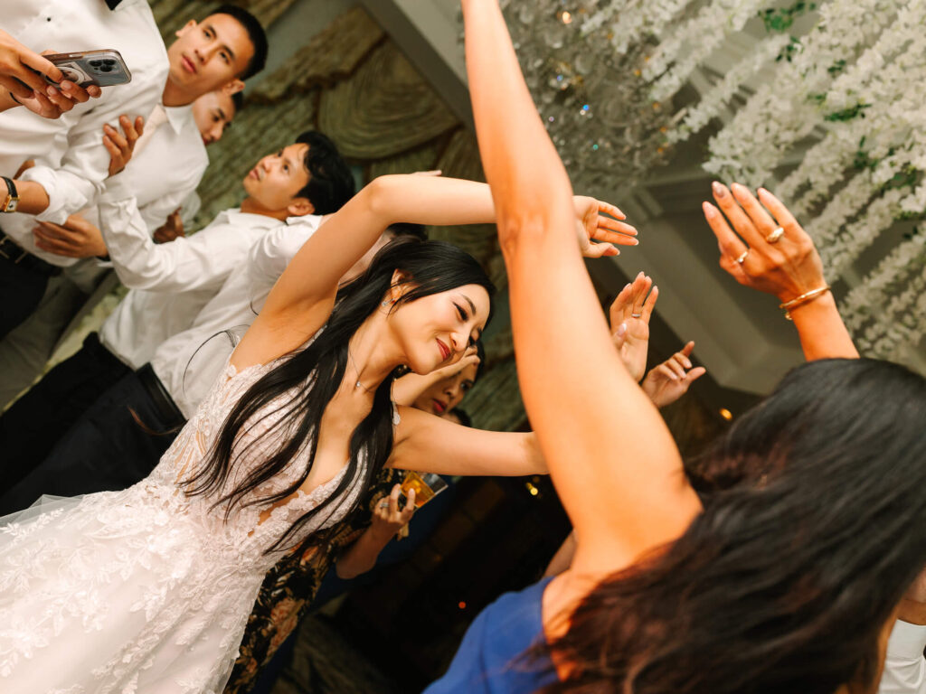 A group of people joyfully dance, with a smiling woman in a white dress taking center stage. Her hair flows as others raise their arms.