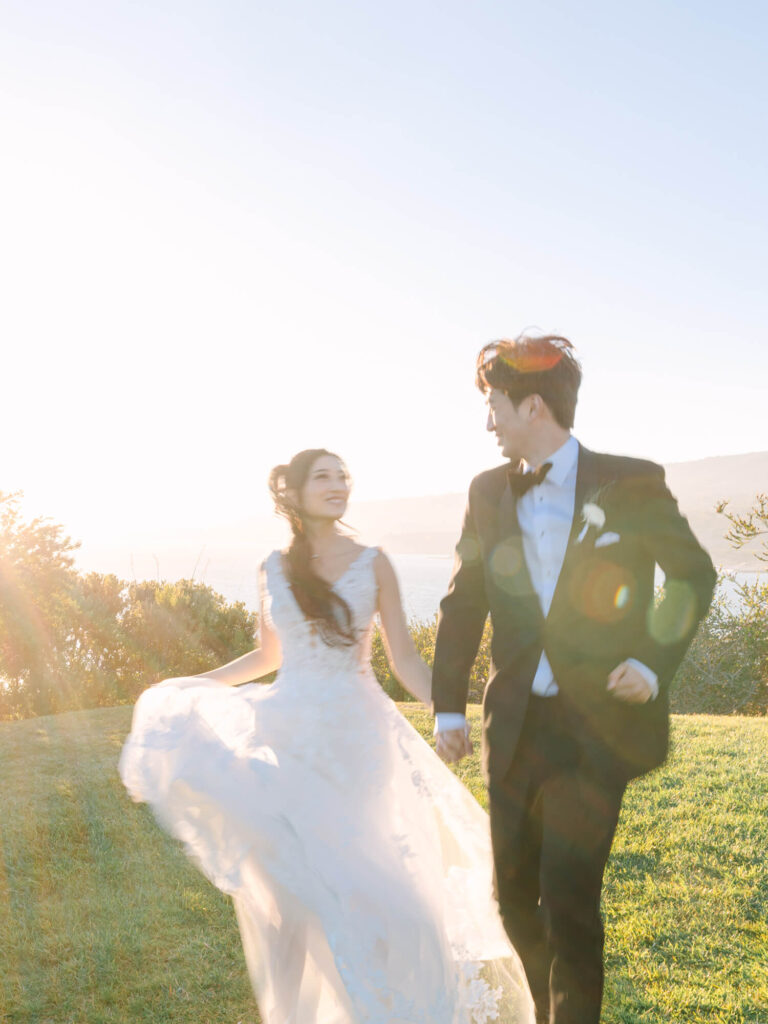 Bride and groom hold hands, joyfully walking on sunlit grass.