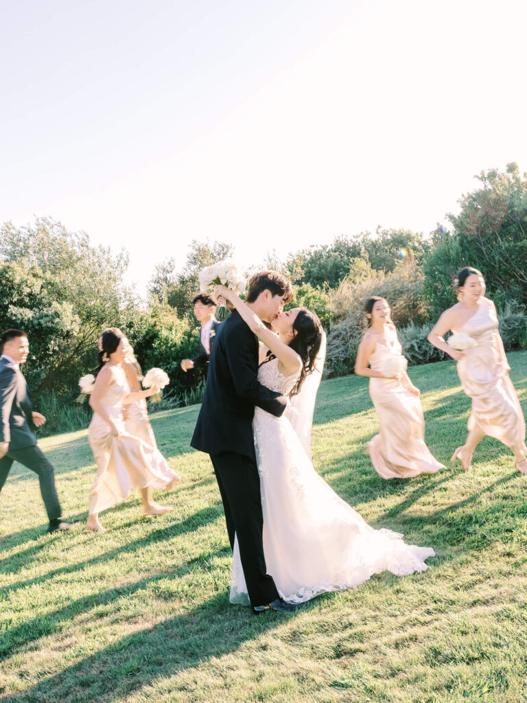 A bride and groom share a joyful kiss in a sunlit field, surrounded by bridesmaids and groomsmen in motion, creating a lively and celebratory scene.