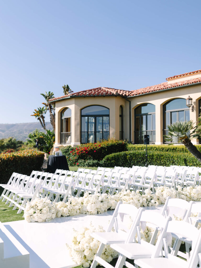 Outdoor wedding venue with white chairs, flower arrangements, and manicured hedges. A beige building with arched windows is in the background under a clear blue sky.